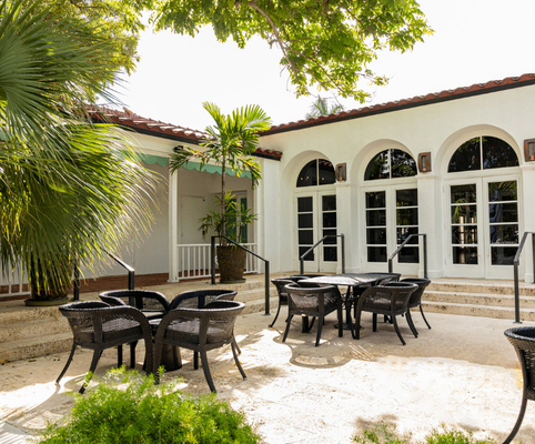 Outdoor patio area with black wicker tables and chairs, surrounded by lush greenery and palm trees, in front of a white building with arched windows and a red-tiled roof.